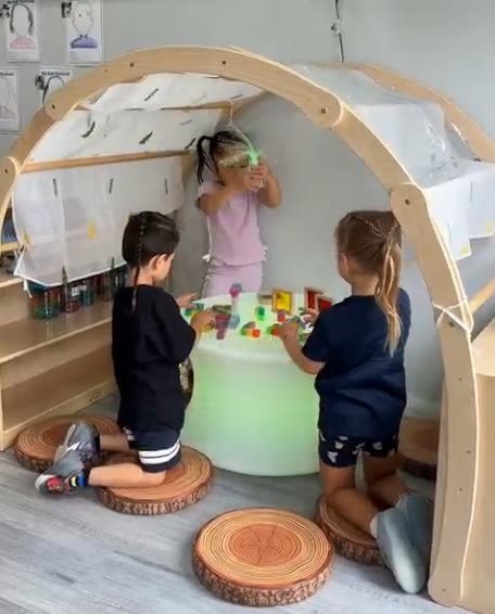 Three children building with translucent blocks on a glowing light table inside a wooden play arch.